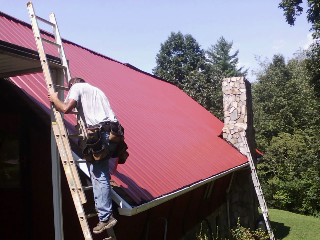 Roofer on a ladder beside a red metal roof and stone chimney — Soffit & Fascia Repair by Roofing Brothers in Foster
